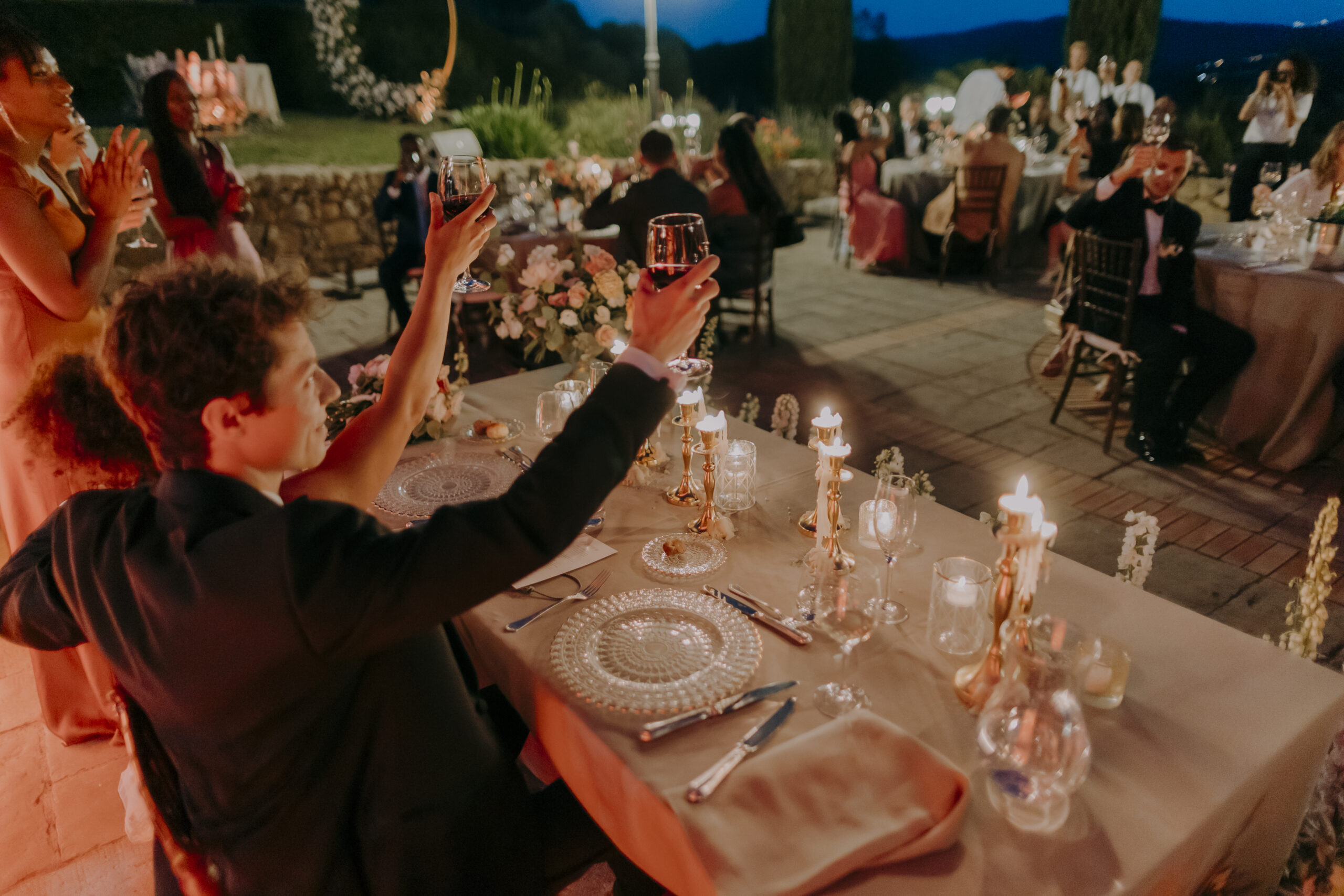Wedding guests raising their glasses during an outdoor candlelit reception.